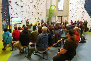 Knapp 100 Personen sitzen auf Bierbänken in einer Kletterhalle und hören einen Vortrag.